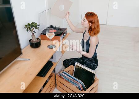 Jeune femme sélectionnant le disque en vinyle dans un conteneur en bois tout en étant assise contre la plaque tournante d'époque dans la chambre Banque D'Images