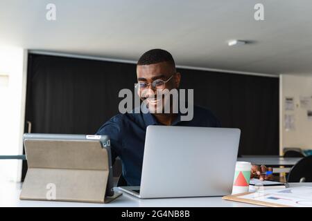 Homme afro-américain adulte positif en vêtements et lunettes de vue habillés à table tout en ayant la vidéoconférence sur tablette près de netbook et de documents wi Banque D'Images