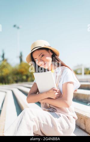 Contenu asiatique femme en chapeau avec livre regardant l'appareil photo tout en se reposant sur les escaliers urbains dans la lumière du soleil Banque D'Images