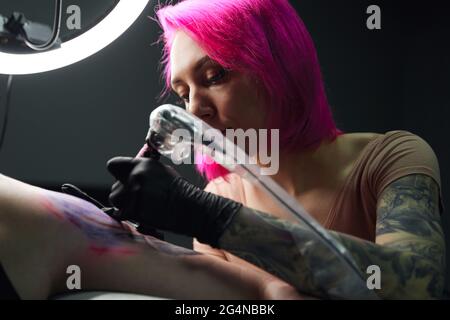 Maître de tatouage sérieux avec cheveux roses dans les gants en utilisant la machine de tatouage professionnelle tout en faisant le tatouage sur l'épaule du client dans le salon de tatouage moderne Banque D'Images