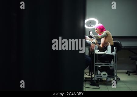 Maître de tatouage sérieux avec cheveux roses dans les gants en utilisant la machine de tatouage professionnelle tout en faisant le tatouage sur l'épaule du client dans le salon de tatouage moderne Banque D'Images