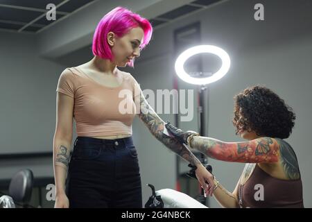 Tatoueur de vue latérale avec gants en latex essuyant un tatouage fraîchement préparé sur le bras de la cliente avec une serviette de table pendant le travail en salon Banque D'Images
