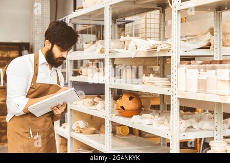 Jeune homme d'affaires séduisant un potter avec une barbe et une moustache travaille dans son atelier. Conserve les enregistrements et transcrites dans un ordinateur portable en inspectant Banque D'Images