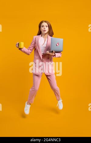 REDHEAD femme saut en utilisant ordinateur portable web projet isolé sur fond jaune en studio, tenant une tasse de café en mains, manquer la date limite, hur Banque D'Images