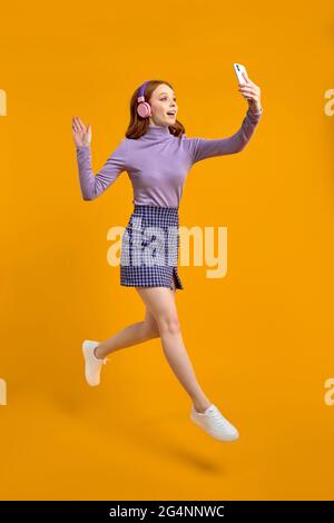 Femme à tête rouge excitée à Heaphones saut, marche et parler au téléphone en ligne via caméra, dire Bonjour, sourire, isolé sur jaune Banque D'Images