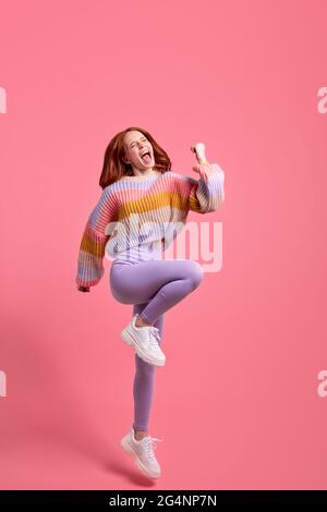 Photo pleine grandeur de heureux rouge cheveux jeune femme hurle oui chanceux gagner les poings hausser porter ensemble décontracté isolé sur fond de couleur rose, réjouissez-vous ici Banque D'Images