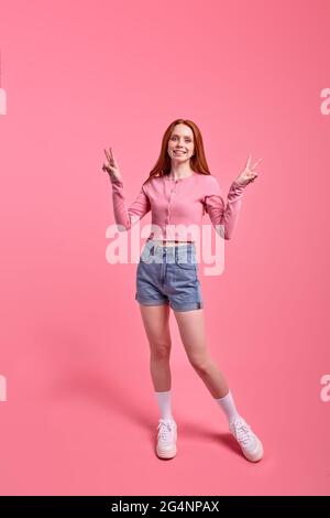 Jolie femme à tête rouge posant, montrant le V-signe isolé sur fond rose vif, portrait de studio. Jeune femme en chemise rose décontractée et Jean denim Banque D'Images