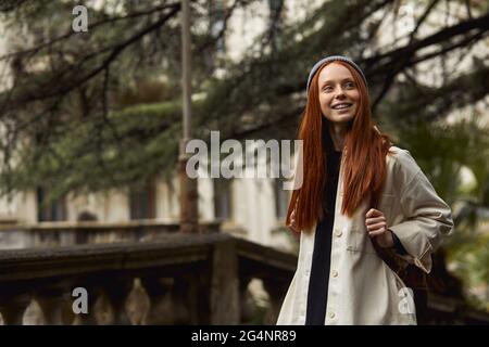 Femme souriante à la tête rouge en manteau à la recherche d'un bel endroit historique, regardant à côté, à des attractions. Les jeunes femmes du caucase passent du temps à l'extérieur, wa Banque D'Images