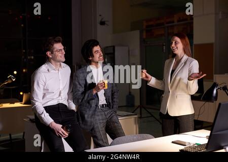 Vue latérale sur les employés de bureau amicaux, les collègues qui parlent après une journée de travail difficile, la nuit. REDHEAD femme et deux gars en tenue formelle discutant, t Banque D'Images