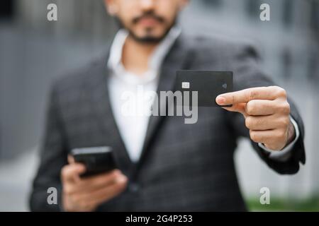 Gros plan de l'homme musulman dans des vêtements formels debout dans la rue de la ville et montrant la carte de crédit noire. Homme occupé utilisant un téléphone cellulaire moderne pour faire des achats en ligne à l'extérieur. Banque D'Images