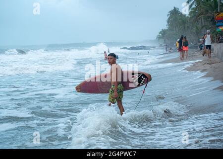 Palomino, Dibulla, la Guajira, Colombie - Mai 24 2021: Jeune Latin Guy marchant avec son surf sur la mer au coucher du soleil sous un ciel nuageux Banque D'Images