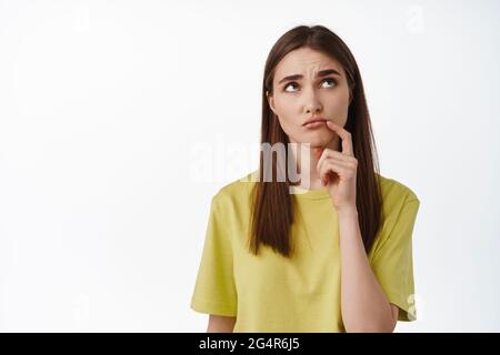 Que faire HMM ? Une jeune femme attentionnée se penche et réfléchit, fait le choix, essaie de décider, pense quoi commander à emporter, se tient contre le blanc Banque D'Images