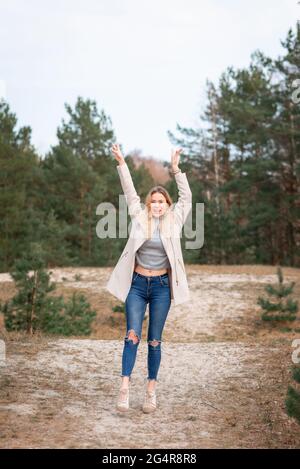 Jeune belle blonde longue cheveux femme debout vue de face avec les mains levées sur fond vert naturel extérieur. Jeune femme bras élevés appréciant Banque D'Images