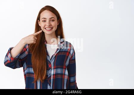 Blanchiment et stomatologie. Souriant redhead jeune fille pointant vers ses dents blanches parfaites, recommander clinique dentaire, debout dans une chemise à carreaux contre Banque D'Images