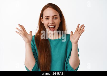 Image de la belle fille heureuse avec des cheveux rouges, des mains de clap et rire joyeuse, appréciant regarder drôle et amusant événement, debout sur fond blanc Banque D'Images