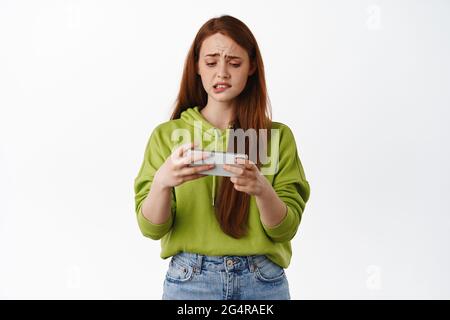 Une fille nerveuse regarde l'écran horizontal du smartphone et mord la lèvre intense, essayant de gagner dans le jeu vidéo mobile, debout dans une tenue décontractée contre blanc Banque D'Images