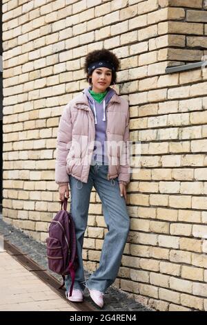 Femme afro-américaine avec sac à dos regardant l'appareil photo près du mur de brique Banque D'Images