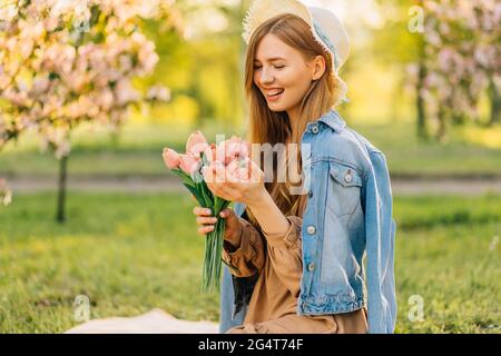 Belle jeune femme dans un chapeau, avec un bouquet de fleurs de printemps, dans le jardin de cerisiers en fleurs un jour de printemps, les pétales de fleurs tombent d'un arbre, printemps Banque D'Images