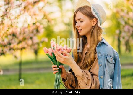 Belle jeune femme dans un chapeau, avec un bouquet de fleurs de printemps, dans le jardin de cerisiers en fleurs un jour de printemps, les pétales de fleurs tombent d'un arbre, printemps Banque D'Images