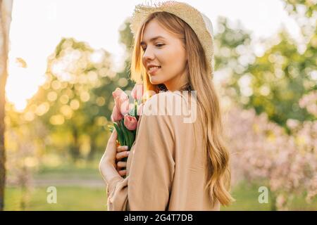 Jolie jeune femme, dans un chapeau de paille d'été, avec un bouquet de tulipes roses, dans un parc de printemps, appréciant la belle nature Banque D'Images