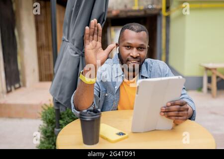 Jeune homme millénaire afro-américain noir souriant assis dans un café à l'intérieur, regardant la caméra et saluant en parlant avec des collègues ou des amis sur vidéo Banque D'Images