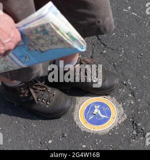 Un marcheur, tenant une copie d'une carte, marche au-dessus d'un marqueur de route pour le sentier côtier de l'île d'Anglesey. Banque D'Images