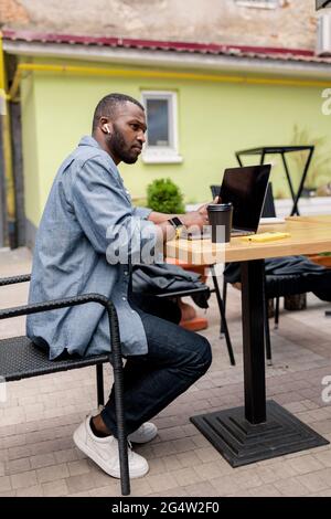 Vue latérale d'un travailleur indépendant afro-américain spécialisé portant des écouteurs sans fil, tenant une réunion en ligne et regardant des cours éducatifs sur un ordinateur portable Banque D'Images