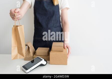 Jeune femme portant un tablier en denim avec les bras croisés debout près de la table avec des boîtes en carton, des emballages en carton et un terminal de paiement moderne. Petit b Banque D'Images