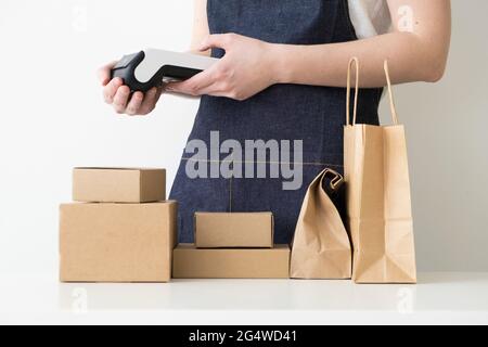 Jeune femme portant un tablier en denim avec les bras croisés debout près de la table avec des boîtes en carton, des emballages en carton et un terminal de paiement moderne. Petit b Banque D'Images