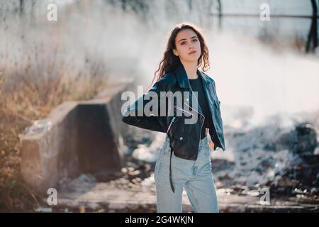 Portrait extérieur d'une belle jeune fille brune dans la veste en cuir noir avec de la fumée en arrière-plan Banque D'Images