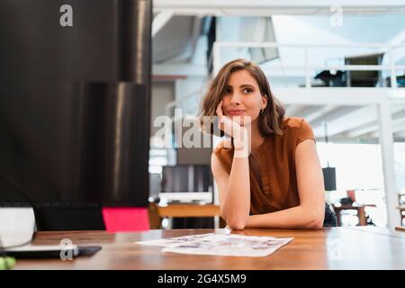 Femme entrepreneure attentive penchée sur le bureau avec la main sur le menton dans le bureau créatif en regardant loin Banque D'Images