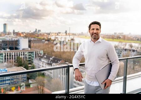 Homme d'affaires souriant avec ordinateur portable penché sur la rampe au balcon dans le bureau Banque D'Images