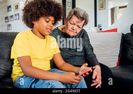 Femme âgée regardant son petit-fils à l'aide d'un smartphone tout en étant assise sur un canapé dans le salon Banque D'Images
