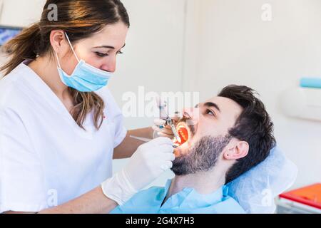 Dentiste féminin portant un masque facial de protection examinant les dents du patient masculin à la clinique médicale Banque D'Images