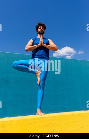 Homme avec les mains claspéd pratiquant Vrikshasana tout en se tenant sur le mur de retenue Banque D'Images