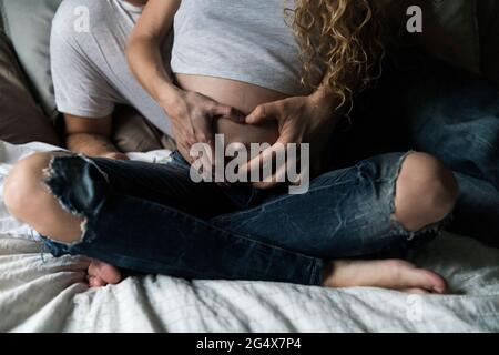 Femme enceinte faisant le signe de la main de forme de coeur sur l'abdomen tout en étant assise avec l'homme dans le lit Banque D'Images