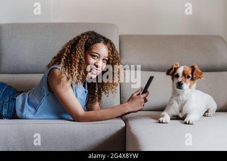 Femme aux cheveux bouclés avec un smartphone allongé sur un canapé près d'un chien dans le salon Banque D'Images