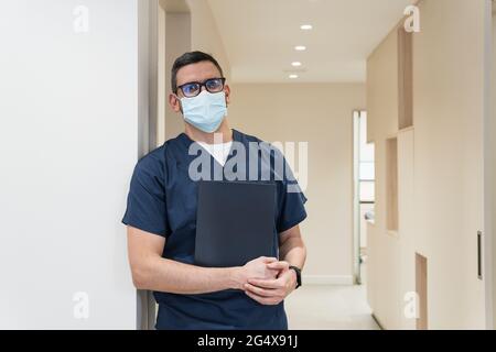 Un dentiste de sexe masculin portant un masque facial de protection debout avec un dossier à la clinique Banque D'Images