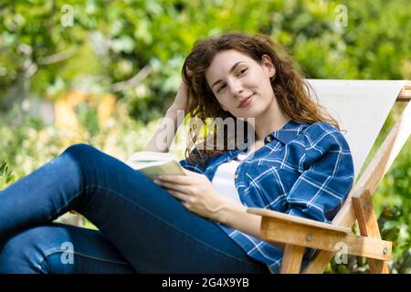 Belle femme avec main dans le livre de cheveux tout en se relaxant sur une chaise dans le jardin Banque D'Images