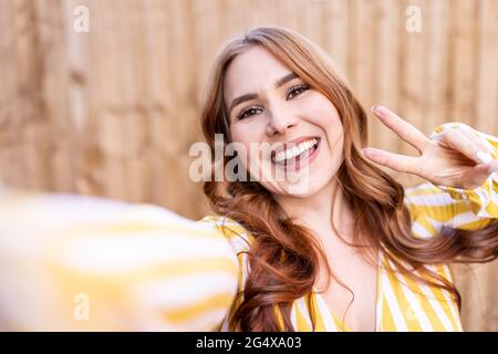 Joyeuse femme gestante signe de paix tout en se tenant contre le mur en bois Banque D'Images