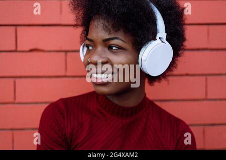 Femme afro souriante avec casque qui regarde loin du mur de briques Banque D'Images