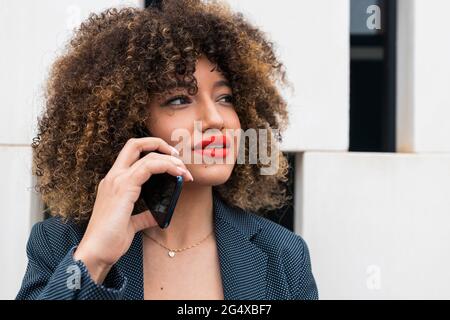 Femme d'affaires afro contemplant tout en parlant sur un smartphone devant le mur Banque D'Images