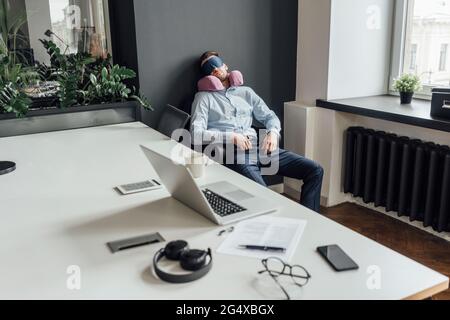 Entrepreneur de sexe masculin fatigué avec oreiller de cou et masque pour les yeux qui se nappent au bureau Banque D'Images