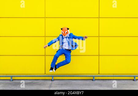 Homme portant un costume bleu vif et un masque tigre sautant contre un mur jaune Banque D'Images