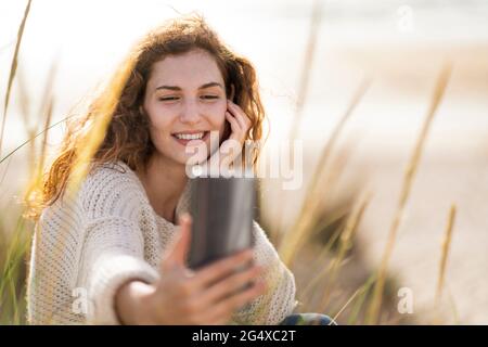 Femme souriante prenant le selfie à travers le smartphone dans les dunes à la plage pendant la journée ensoleillée Banque D'Images