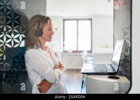 Femme souriante du service clientèle, bras croisés devant l'ordinateur portable au bureau Banque D'Images