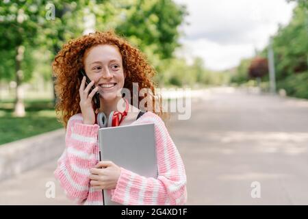 Bonne femme à tête rouge avec des conversations mauriquement sur un téléphone intelligent au parc tout en regardant loin Banque D'Images