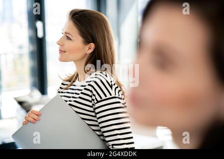 Femme d'affaires souriante avec un ordinateur portable qui envisage de se tenir debout au bureau Banque D'Images