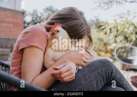 Femme embrassant le poulet tout en étant assise sur une chaise dans l'arrière-cour Banque D'Images