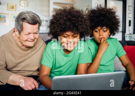 Garçon souriant utilisant un ordinateur portable tout en étant assis par une grande grand-mère et un frère dans le salon Banque D'Images
